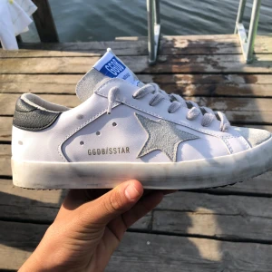 Golden Goose vita sneakers med stjärna - Golden Goose sneakers i vitt skinn med grå mockadetaljer och ikonisk stjärna på sidan. Grå snörning, svart hälparti med guldfärgad logga och slitna detaljer på sulan för en cool, vintage look. Perfekta för dig som gillar streetstyle och unika detaljer.