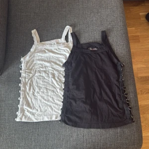 Suzette ribbade linnetoppar med ringdetaljer - Två ribbade linnetoppar från Suzette, en i svart och en i vitt. Båda har breda axelband och coola silverringar längs sidorna som ger en edgy vibe. Materialet är mjukt och stretchigt, perfekt för att styla med jeans eller kjol. Säljes tillsammans!