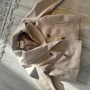 Beige kappa från Vila, stl 34 - Stilren beige skepparkavaj från Vila med dubbelknäppning och klassisk krage. Jackan har raka linjer, två fickor framtill och är helfodrad. Perfekt för dig som gillar en clean och tidlös look. Materialet är mjukt och känns lyxigt. Använd 1 gång. Nyskick