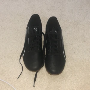 Svarta fotbollsskor från Puma - Snygga svarta fotbollsskor från Puma med vita detaljer och klassisk logga på tungan. Skorna har snörning, låg profil och en platt sula i brunt gummi. Ovandelen är i syntetmaterial och designen är sportig och stilren.