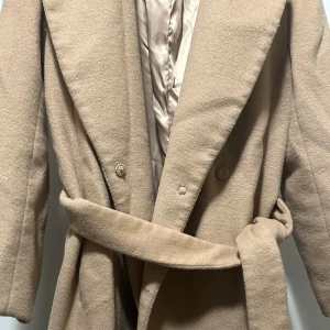 Beige kappa med bälte och knappar - Stilren beige kappa med bred krage, knappar framtill och ett matchande bälte i midjan. Yttermaterialet är mjukt och känns som ull, medan fodret är slätt och glansigt. Perfekt för dig som vill ha en klassisk och snygg look under höst och vinter.