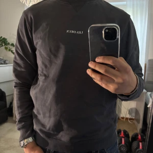 Mörkgrå sweatshirt från Les Deux - Snygg mörkgrå sweatshirt från Les Deux med vit broderad logga på bröstet. Tröjan har rund halsringning, ribbade muddar och är tillverkad i mjuk bomull. Perfekt för dig som gillar stilren och clean look.