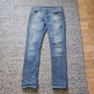 Blå jeans från Nudie Jeans Co - Säljer ett par klassiska blå jeans från Nudie Jeans Co med snygga slitningar och kontrastsömmar. Jeansen har raka ben, normal passform och coola broderade bakfickor. Perfekta för dig som gillar en tidlös och avslappnad stil. Jag har bytt översta knappen litet märke i tyget. Hör av er vid minsta fundering🙏