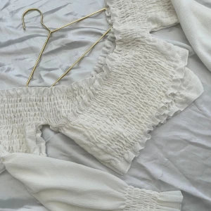 Vit smockad blus med volang - Supersöt vit blus med smockad kropp och volangdetaljer längs kanterna. Toppen har långa puffärmar med smock vid handlederna och en offshoulder-design.