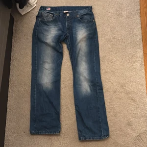 True Religion blå jeans med raka ben - Snygga blå jeans från True Religion med raka ben och coola blekningar framtill och baktill. Jeansen har klassiska kontrastsömmar, fem fickor och tydlig logga med döskalle över den bakre bakfickan. Skriv till mig privat för mer detaljer