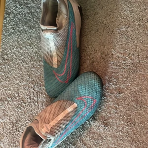 Nike phanton blå fotbollsskor strl 40,5 - Säljer ett par Nike phantom fotbollsskor i blått med rosa swoosh och vita detaljer. Skorna har en strömlinjeformad design med täckt snörning och svart dobbsula för bästa grepp på planen. Ovandelen är i syntetmaterial som ger lätt känsla och bra bollkontroll. Dom är i fint skick och är endast använda en säsong. 