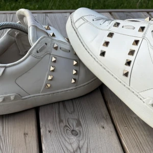 Valentino Rockstud vita sneakers - Snygga vita Valentino Rockstud sneakers i skinn med ikoniska guldiga nitar längs sidorna och på tungan. Klassisk låg modell med snörning och rund tå. Perfekta för dig som vill sticka ut med en lyxig vibe. Skick 8,5/10 lite creasningar bara. Skriv gärna vid frågor. Storlek 41 men passar lite större. Priset är inte hugget i sten