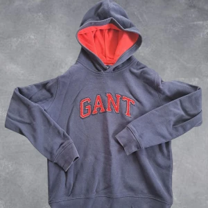 GANT hoodie - Säljer en blå hoodie från GANT med röd insida i huvan och broderad logga i rött och vitt på bröstet. Klassisk modell med långa ärmar och mjukt material, perfekt för en avslappnad stil.