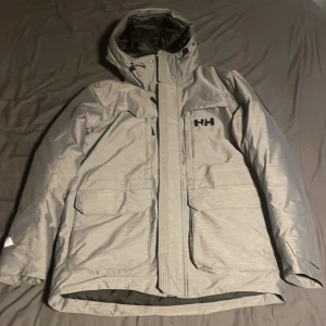 Grå dunjacka från Helly Hansen - Snygg grå dunjacka från Helly Hansen med svart innerfoder, stor huva och praktiska fickor framtill. Jackan har dragkedja och är perfekt för kalla dagar. Klassisk design med HH-logga på bröstet och ryggen. Passar dig som vill ha både stil och funktion. Skicket på den är som ny då den används extremt få gånger.