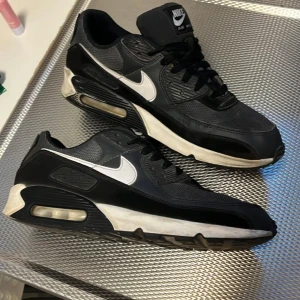 Nike Air Max 90 svart/vit  - Säljer ett par klassiska Nike Air Max 90 sneakers i svart med vita detaljer och sula. Skorna har en ikonisk siluett, synlig Air Max-dämpning och är tillverkade i en mix av skinn och syntetmaterial. Perfekta för dig som gillar streetstyle och sportig vibe.