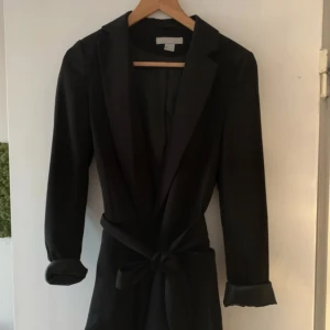Svart kappa från H&M (snöret är borta!) - Stilren svart kavaj från H&M i storlek 34. Kavajen har klassisk krage, långa ärmar och ett snyggt bälte i midjan som knyts framtill. Ärmsluten är uppvikta med blank insida för en cool kontrast. Perfekt för dig som gillar en clean och modern look.