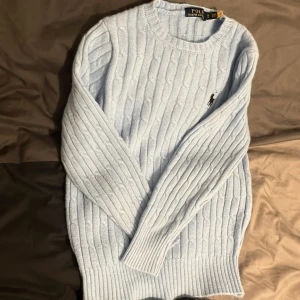 Ljusblå kabelstickad tröja Polo Ralph Lauren - Kabelstickad ljusblå tröja från Polo Ralph Lauren med rund halsringning och broderad logga på bröstet. Tröjan har långa ärmar och ribbade muddar vid ärmslut och nederkant. Perfekt för lager-på-lager och snygg till jeans. Storlek M men passar även s 
