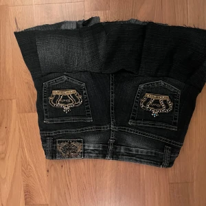 Svart jeanskjol med guldiga detaljer - Cool svart jeanskjol med rå kant och volang nertill. Kjolen har snygga guldiga broderier och dekorativa detaljer på både fram- och bakfickor. Flera knappar framtill och slitna kanter ger en edgy vibe. Perfekt för dig som vill sticka ut med din stil.😊