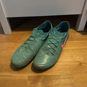 Nike Phantom GT fotbollsskor turkos - Nike Phantom GT fotbollsskor i turkos med rosa Swoosh och blå detaljer. Skorna har snörning, rund tå och är tillverkade i syntetmaterial med stickad känsla för extra bra bollkontroll. Perfekta för dig som vill sticka ut på planen. Vid några funderingar hör av dig // William 