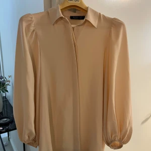 Beige skjorta från REFKA med puffärm - Stilren beige skjorta med klassisk krage och dolda knappar framtill. Skjortan har långa puffärmar med resår vid ärmslut och är tillverkad i ett mjukt, slätt material. Perfekt för dig som gillar en clean och elegant look.