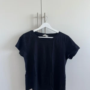 Svart basic t-shirt i bomull - Enkel svart t-shirt med rund hals och korta ärmar. Modellen är klassisk och passar till allt, tillverkad i mjuk bomull för en skön känsla. Perfekt att matcha med jeans eller shorts för en clean look.
