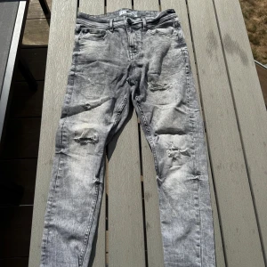 Grå slitna skinny jeans 28/30 - Säljer ett par grå skinny jeans med slitningar och hål på knäna. Jeansen har en tvättad look, fem fickor och klassisk knappgylf. Perfekta för dig som gillar en lite ruffare stil och vill ha ett par bekväma jeans med attityd.