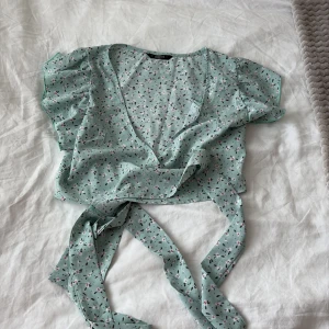 Blommig omlottopp från SHEIN - Supersöt ljusgrön omlottopp från SHEIN med blommigt mönster i rosa och vitt. Toppen har korta volangärmar och knyts i midjan för en snygg passform. Perfekt för vår och sommar när du vill ha en lätt och luftig look.