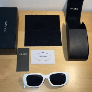 Vita solglasögon från Prada - Snygga solglasögon från Prada med vit, kantig båge och svarta glas. Modellen har en modern, chunky design och tydlig Prada-logga på sidan. Kommer med originalfodral, putsduk och äkthetskort. Perfekt accessoar för att lyfta din stil.