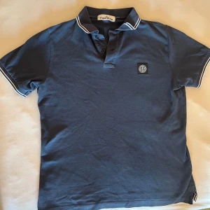 Blå Stone Island pikétröja - Snygg marinblå pikétröja från Stone Island med klassisk krage och vita ränder på krage och ärmslut. Tröjan har korta ärmar, knappslå framtill och ikonisk logga på bröstet. Tillverkad i bomull för en skön känsla och stilren look.                             Passar small/ medium