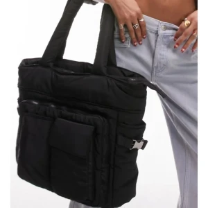 Helt ny Svart stor axelväska med fickor - En svart, rymlig axelväska med flera praktiska ytterfickor och breda handtag. Väskan har dragkedja upptill och spänndetaljer på sidorna. Perfekt för dig som vill ha plats för allt och gillar en clean, urban stil.