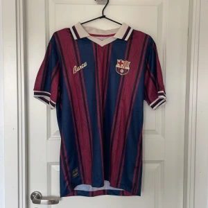 Barça retro fotbollströja randig - Snygg retroinspirerad FC Barcelona fotbollströja med vertikala blå och vinröda ränder, klassisk vit krage med blå och vinröda detaljer. Broderad Barça-text och klubbmärke på bröstet. Tillverkad i lätt och ventilerande polyester, perfekt för match eller samling.