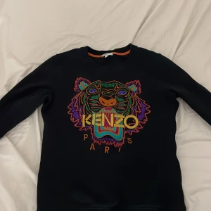Svart Kenzo tröja med tigerbrodyr XS - Svart tröja från Kenzo i storlek XS med ikonisk färgglad tigerbrodyr och Kenzo Paris-text på bröstet. Tröjan har rund halsringning, långa ärmar och är tillverkad i mjukt material. Perfekt statement-plagg för dig som gillar streetstyle.