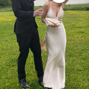 Balklänning/klänning - Elegant långklänning i champagnefärgad satin med djup v-ringning, smala axelband och öppen rygg med knytband. Klänningen har en figurnära passform och faller snyggt längs kroppen. Perfekt för bal eller festliga tillfällen. Använd 1 gång.