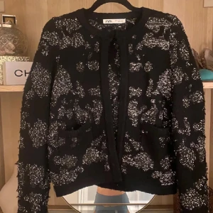 Glittrig svart cardigan från Zara - Svart cardigan från Zara med glittriga detaljer i silver. Koftan har rund halsringning, två fickor framtill och är långärmad. Perfekt för dig som vill sticka ut med en snygg och trendig look. Aldrig använd.