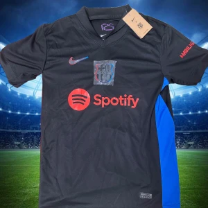 FC Barcelona svart Nike fotbollströja - Snygg svart FC Barcelona fotbollströja från Nike med röd Spotify-logga på bröstet och klubbmärke. Blå detalj på sidan och UNHCR-tryck på ryggen. Kortärmad modell i lätt och ventilerande material, perfekt för match eller träning.