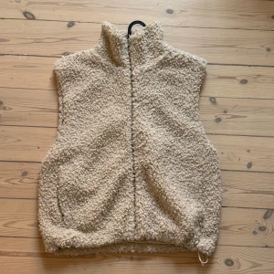Beige teddyväst från H&M - Mysig beige teddyväst från H&M med hög krage och dragkedja framtill. Västen är i fluffigt syntetmaterial och har en loose passform, perfekt att slänga över en hoodie eller stickad tröja. Fickor på sidorna och justerbar dragsko nertill för extra komfort🩷🌸🌻