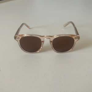 Beige solglasögon från John Henric - Snygga solglasögon från John Henric med transparent beige båge och mörka runda glas. Kommer med originalfodral och mjuk påse. Perfekta för soliga dagar och ger en clean och modern vibe.