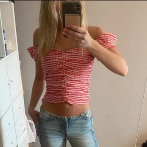 Rutig röd offshoulder croppad topp - Superfin croppad topp med rödvitt rutigt mönster och offshoulder-ärmar. Toppen har smockad passform och rynkade detaljer framtill som ger en snygg siluett. Perfekt för varma dagar och en trendig look.