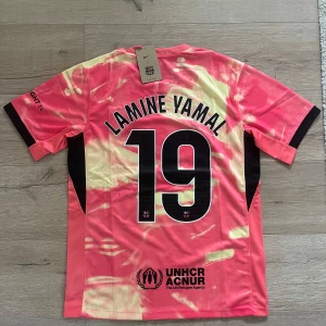 Barcelona Lamine Yamal 19 Nike fotbollströja - FC Barcelona fotbollströja med Lamine Yamal 19-tryck. Tröjan är kortärmad och har ett unikt rosa och ljusgult mönster med svarta detaljer. Nike-logga, FCB-märke, Spotify och UNHCR-loggor. Tillverkad i lätt och ventilerande polyester.
