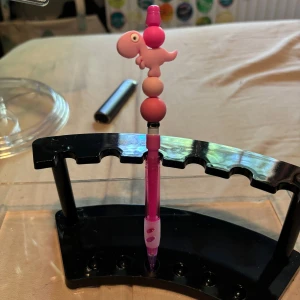 Stift penna - Bilderna visar en rosa penna med ett sött dinosaurie- och pärldekor på toppen, placerad i ett svart pennställ. 