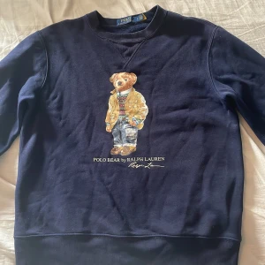 Mörkblå Polo Bear tröja från Ralph Lauren - Snygg mörkblå sweatshirt från Polo Ralph Lauren med ikoniska Polo Bear-trycket på bröstet. Tröjan har rund hals, långa ärmar och är gjord i mjuk bomull. Perfekt för dig som gillar klassisk streetstyle med en twist.