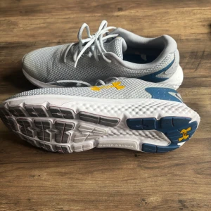 Gråa Under Armour sneakers med mesh - Snygga grå sneakers från Under Armour Charged Rogue med ventilerande mesh-ovandel, vita och blå detaljer samt gult logotryck på sidan. Skorna har snörning och en bekväm, dämpad sula som passar perfekt för träning eller vardagsstil. 