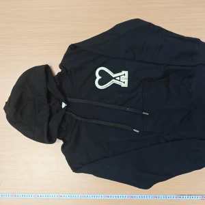 Svart hoodie från Ami med hjärtlogga - Svart hoodie från Ami med stor vit hjärtformad A-logga på bröstet. Klassisk känguruficka, dragsko i huvan och ribbade muddar. Skön och stilren modell i bomull, perfekt för dig som gillar streetwear och vill ha något som sticker ut.