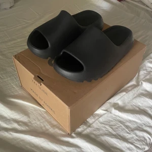 Svarta adidas Yeezy Slide sandaler - Svarta adidas Yeezy Slide sandaler med chunky, räfflad sula och öppen tå. Tillverkade i syntetmaterial för en clean och futuristisk vibe. Perfekta för chill dagar och streetwear-stil. Box medföljer.