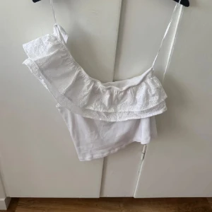 Vit offshoulder topp med volang - Superfin vit topp med offshoulder och volangdetalj. Ena axeln har smalt axelband och den andra är bar, vilket ger en snygg asymmetrisk look. Toppen har broderade spetsdetaljer på volangen och är perfekt för sommardagar.