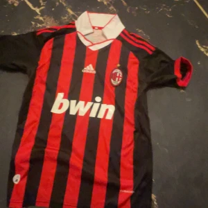 AC Milan Adidas fotbollströja röd/svart - AC Milan fotbollströja från Adidas med klassiska röda och svarta vertikala ränder, vit krage och klubbmärke på bröstet. Tröjan har korta ärmar, Bwin som sponsortryck och är tillverkad i lätt och ventilerande material. Perfekt för dig som älskar italiensk fotboll.
