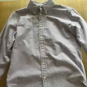 Ljuslila skjorta från Ralph Lauren - Klassisk ljuslila skjorta från Ralph Lauren med button-down krage och broderad logga på bröstet. Skjortan har lång ärm, normal passform och är tillverkad i mjuk bomull. Perfekt för dig som gillar stilrena och tidlösa plagg.