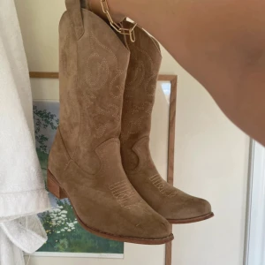 Beiga cowboy boots i mocka - Snygga beiga cowboy boots i mocka med klassiska broderade detaljer och låg klack. Bootsens spetsiga tå och träfärgade sula ger en riktigt trendig westernkänsla. Perfekta för dig som vill sticka ut med en cool och tidlös stil.