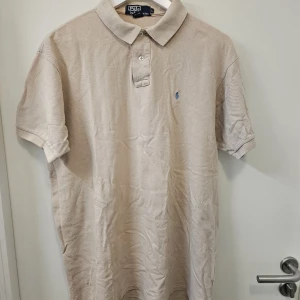 Beige pikétröja från Polo Ralph Lauren made in usa - Säljer en klassisk och tidlös pikétröja från Polo Ralph Lauren. Denna tröja är tillverkad i USA och har den ikoniska Polo-hästen broderad i en snygg blå nyans. En perfekt basplagg för en stilren look! Detaljer: Märke: Polo Ralph Lauren Färg: Ljusbeige / Sand (en mångsidig neutral färg som passar till det mesta) Storlek: L (Large) Skick: Bra begagnat skick med normalt bruksslitage. Färgmatchningen är fortfarande fin och materialet är intakt. Material: 100% bomull -- mjuk och bekväm kvalitet. 