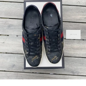 Gucci ace med tiger - Snygga Gucci sneakers i mörkblått med klassiska röda och blå ränder på sidorna. Skorna har ett coolt tigerprint och GG-monogram över hela ovandelen. Baksidan har metallicdetaljer i rött och blått. Perfekta för dig som vill sticka ut med lyxig streetstyle. Ett snöre är av jag har bara inte orkat bytt för att jag endå inte knöt dem men de är enkelt att fixa nya. Kom privat för mer information eller bilder