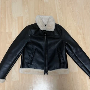 Svart skinnjacka med fuskpäls - Snygg svart skinnjacka med krage och detaljer i beige fuskpäls. Jackan har dragkedja framtill, två fickor och coola spännen vid halsen. Perfekt för kalla dagar när du vill ha en stilren och edgy look.
