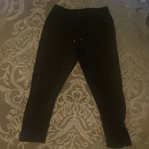 Svarta mjukisbyxor från Nike - Svarta mjukisbyxor från Nike med diskret logga på benet. Byxorna har snörning i midjan och är tillverkade i ett mjukt material som passar perfekt för chill eller träning. Modellen är något croppad och har en relaxed fit.
