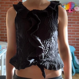Svart volangblus från Gina Tricot - Supersnygg svart blus från Gina Tricot med volangdetalj framtill och crinklad struktur. Ärmlös modell med knyt längst ner och en cool, vågig form. Perfekt för dig som vill sticka ut lite extra med din outfit! Storlek 164 men passar xs-s