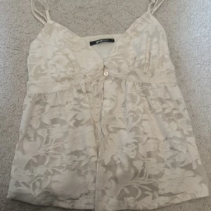 Blommig spetslinnetopp från Gina Tricot - Supersöt ljusbeige linnetopp från Gina Tricot med blommigt spetsmönster och tunna axelband. Toppen har v-ringning och knappar framtill, perfekt för en somrig look. Lätt och luftig känsla med fina detaljer.