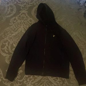 Svart hoodiejacka från Lyle & Scott - Svart hoodiejacka från Lyle & Scott med dragkedja framtill och klassisk gul logga på bröstet. Jackan har lång ärm, huva och är tillverkad i mjukt bomullsmaterial. Perfekt för en chill och stilren look.